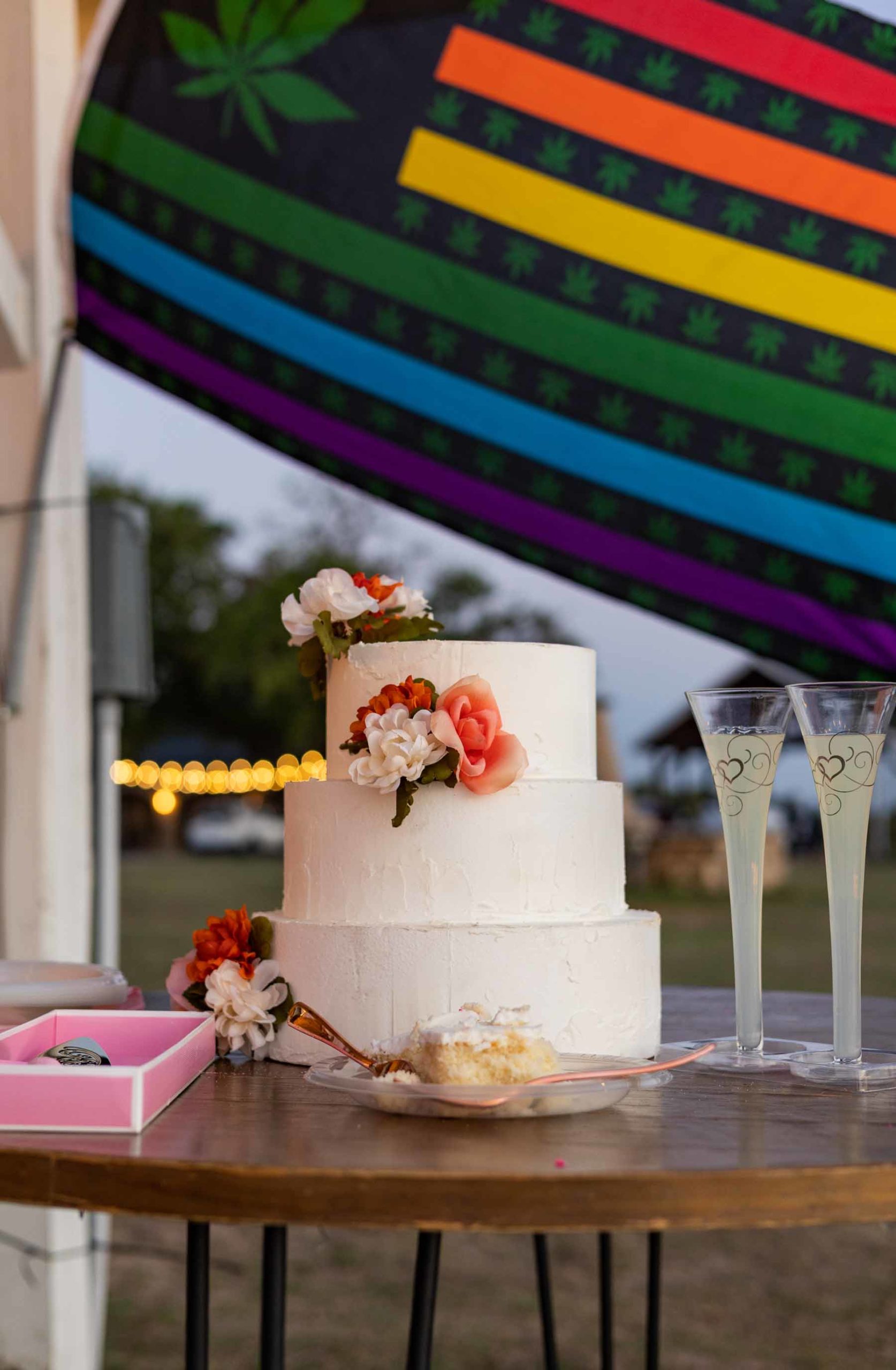 black rainbow pride flag with marijuana plant and a wedding cake topped with flowers, champagne flutes with gold hearts