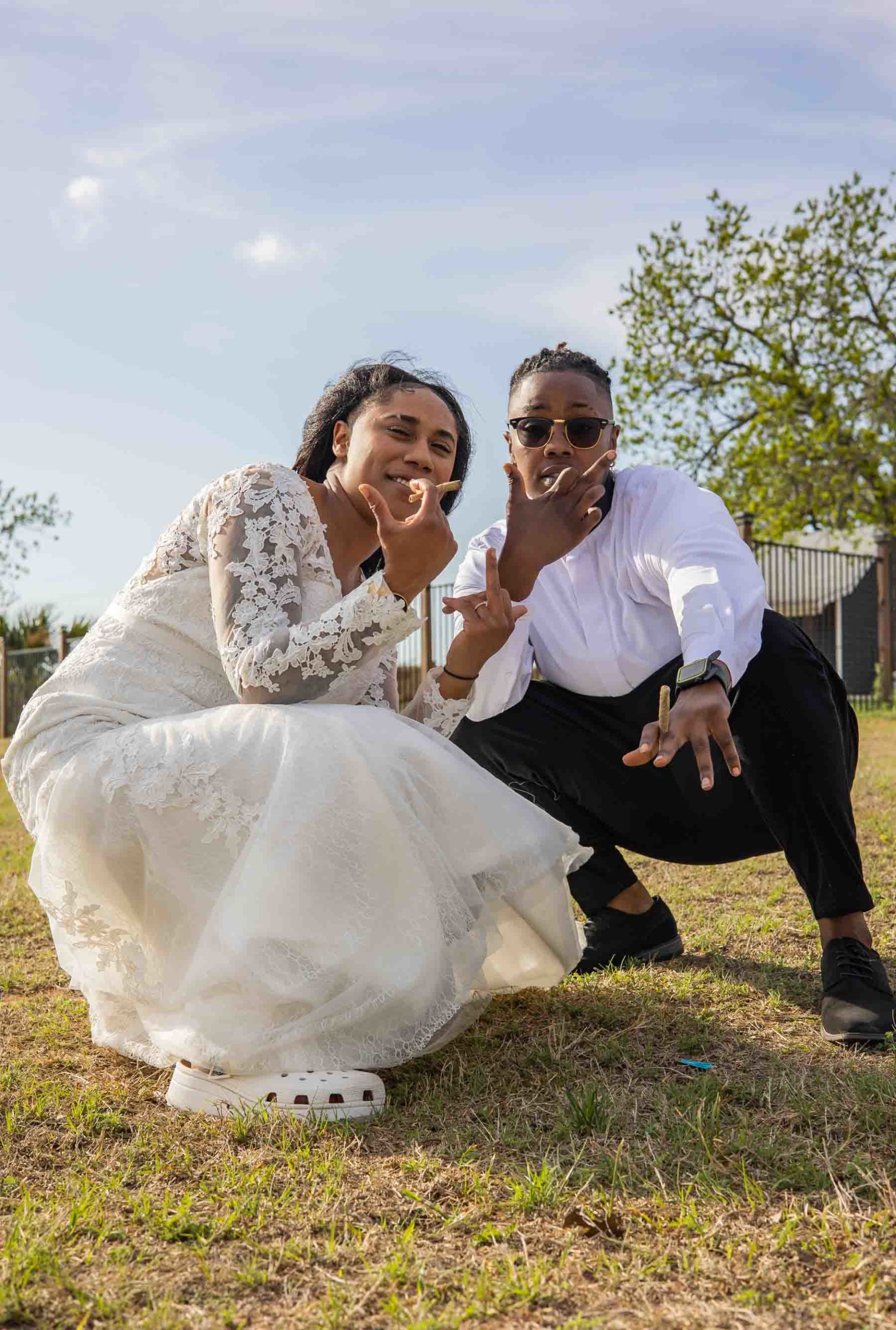 Black lesbian bride and Black wedding officiant squat down holding weed blunts, shooting birds with their fingers and look at camera