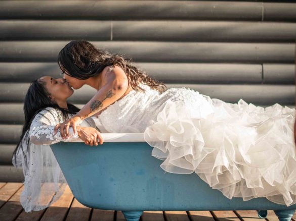 Black lesbian couple laying in blue bathtub, kissing, wearing white wedding gowns