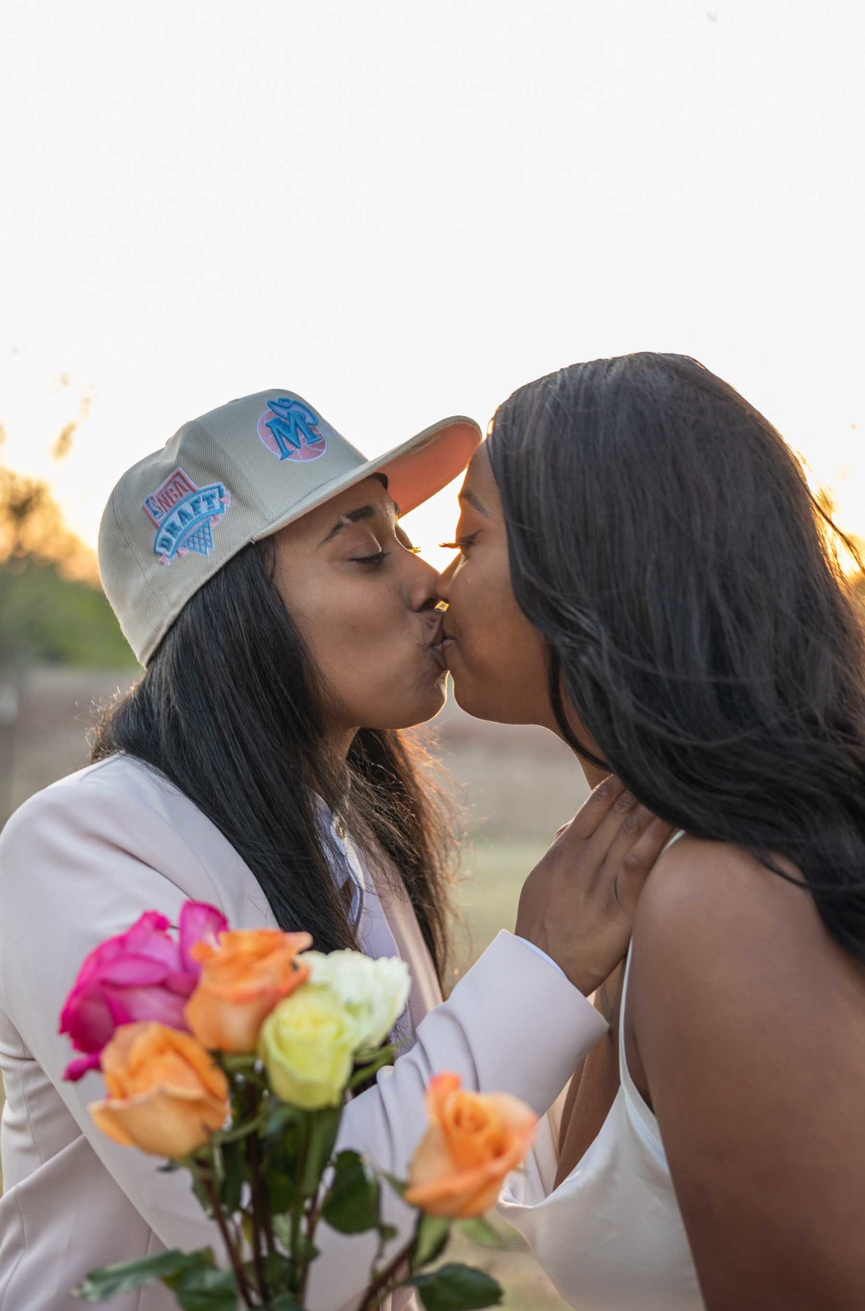 Black lesbian couple kiss, one is in a hat and a pink suit and the other is wearing a silk white dress with thin straps