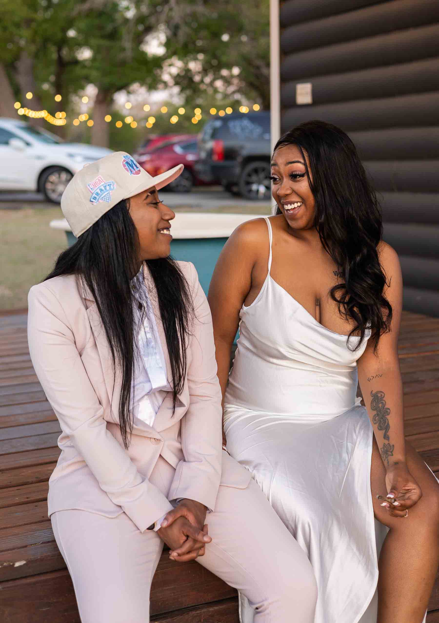 Black lesbian couple smiling at each other, one is in a pink suit and the other is wearing a silk white dress with thin straps