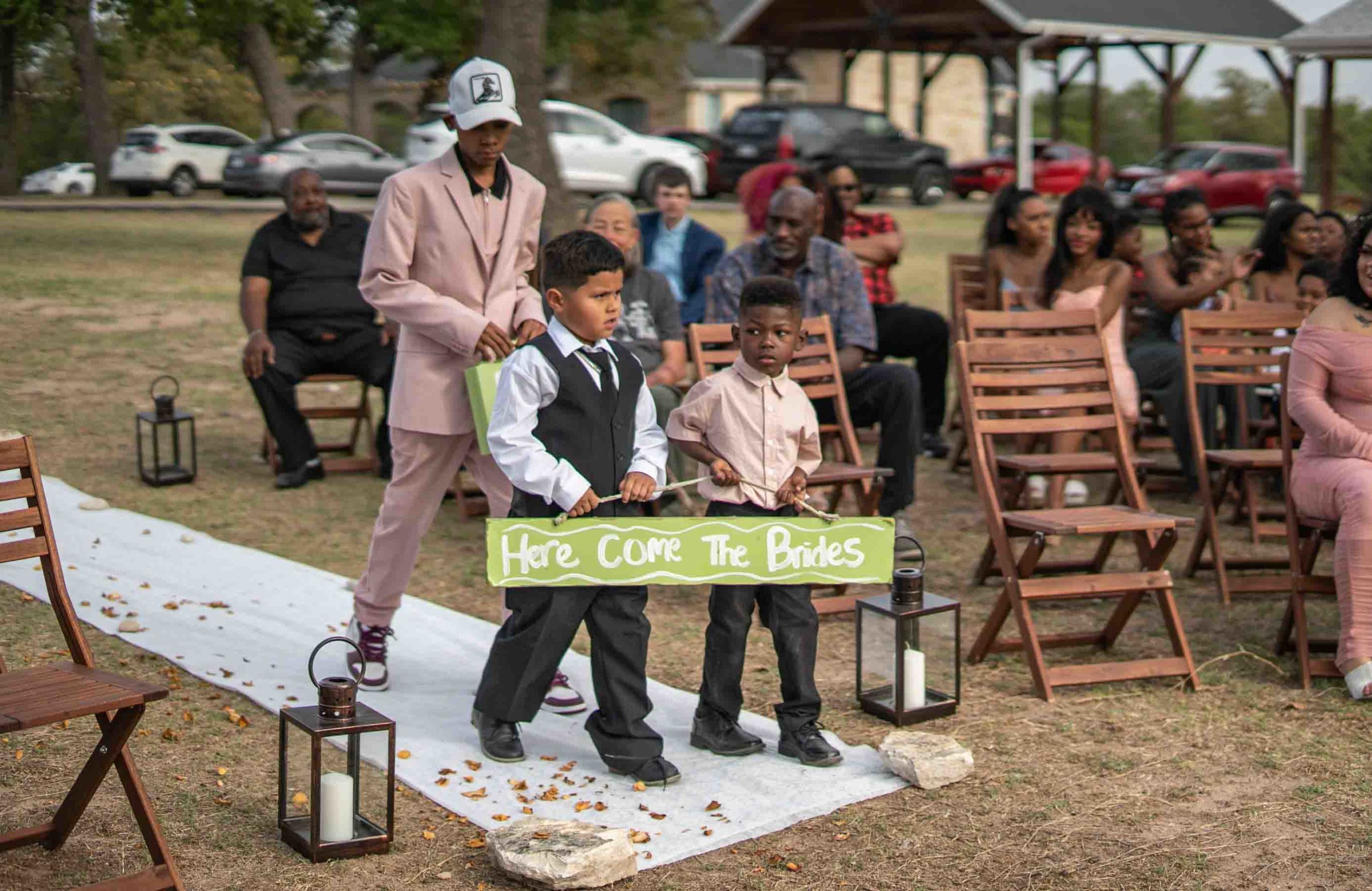 two boys carry a hand painted green sign reading Here Come The Brides followed by a taller boy in a pink suit carrying a green suitcase painted with the words Ring Security while wedding guests watch