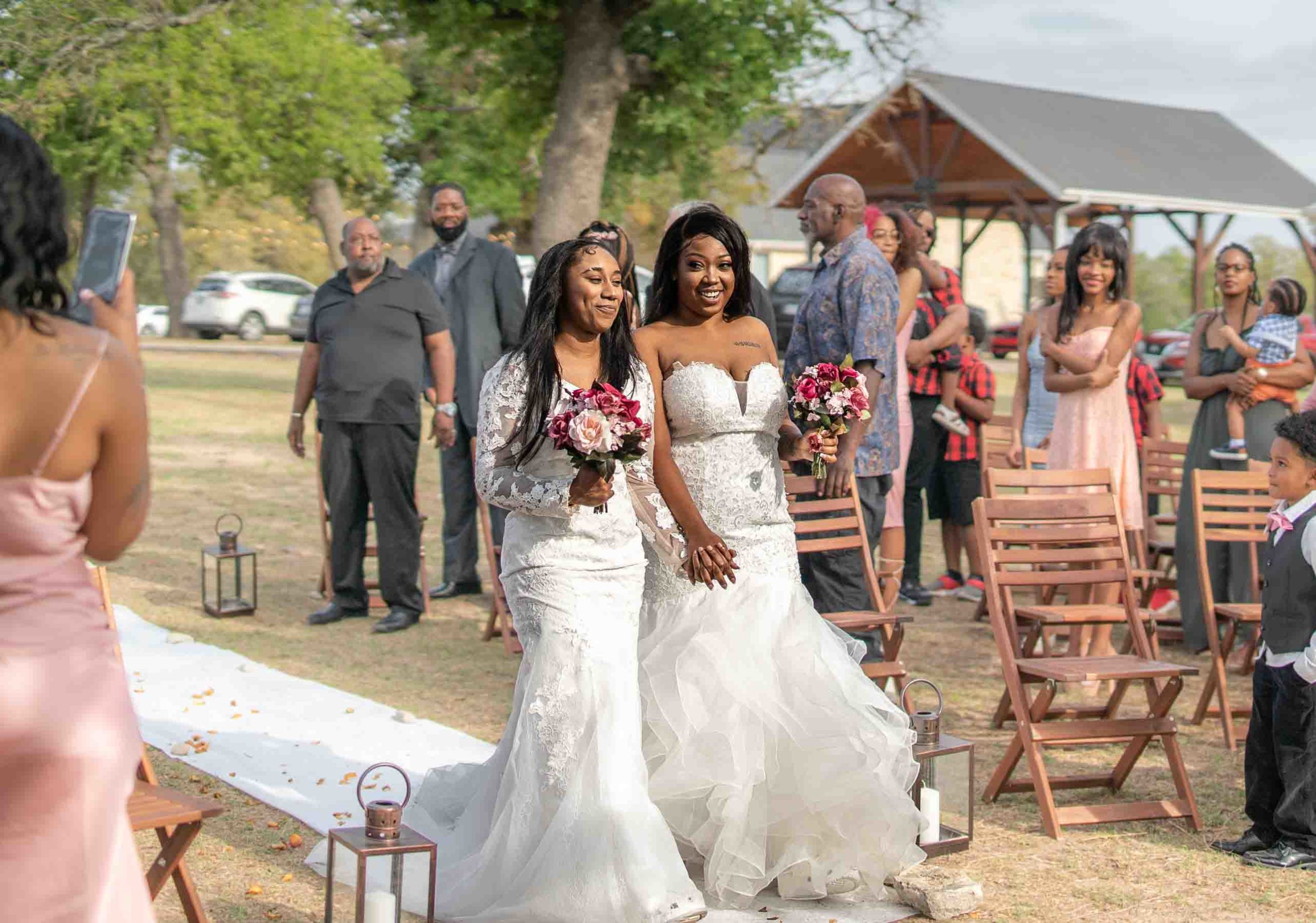 two Black lesbian brides smile as they walk up the aisle together holding hands and bouquets with pink flowers as wedding guests watch