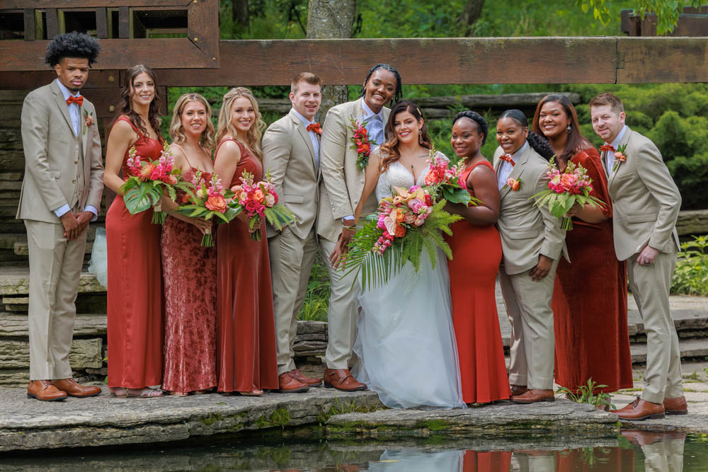 Wedding party of mixed genders and races. The bridesmaids are wearing gowns in shades of burnt orange. Others are wearing tan suits with burnt orange bowties.