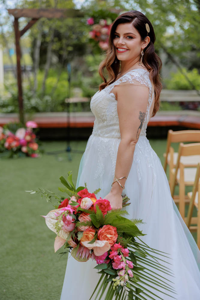 white woman with long brown hair wearing a white wedding gown with capped sleeves smiles and looks at the camera while holding a tropical bouquet of flowers in a shade of pinks and oranges