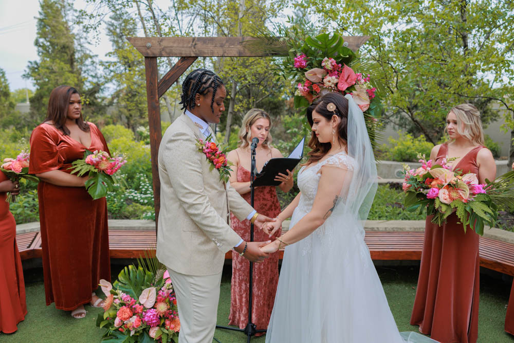 gay wedding with a Black woman in a cream suit and dark orange boutonniere holding hands with a white woman with long dark brown hair in a white veil and white wedding dress with capped sleeves. behind the couple is a wooden wedding arch with a white woman officiant in a dark orange dress. on either side of the marriers are women wearing dark orange gowns and holding vibrant wedding bouquets