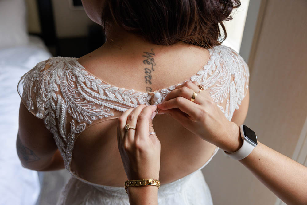 A white woman with dark brown hair's back is showing the back of a wedding gown with an open back being buttoned at top by white hands.