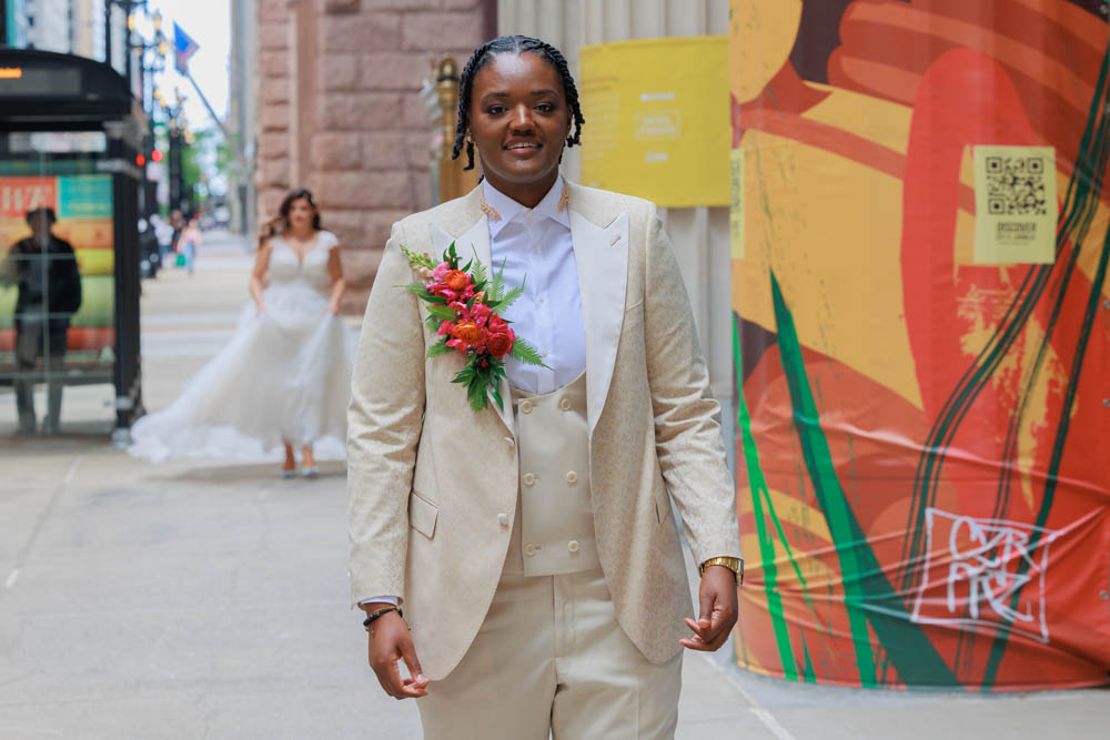 first look on wedding day - a Black woman in a tan suit with black braids and a large red boutonniere stands in foreground while a white bride with long brown hair in a white wedding gown approaches from behind