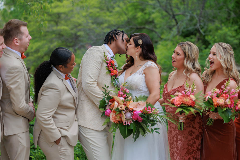 lesbian couple kissing at wedding surrounded by their wedding guests. some are wearing tan suits and some are wearing shades of dark orange and cinnamon