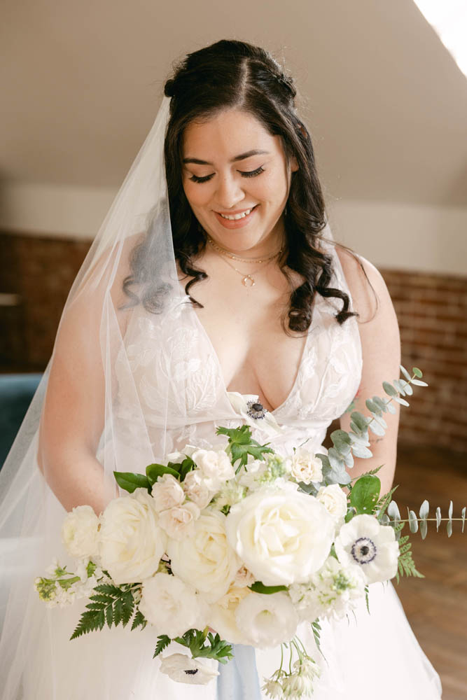 brunette bride with fair skin wearing a white veil and a v-neck white wedding gown holding a wedding bouquet full of a variety of white flowers. she is looking down and smiling.