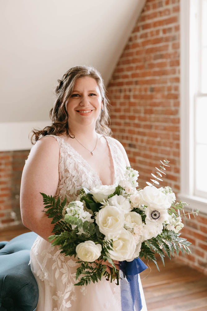 fair-skinned blonde bride holding wedding bouquet of white flowers and blue ribbon. her v-neck wedding dress has a deep plunge. she is looking at the camera and smiling.