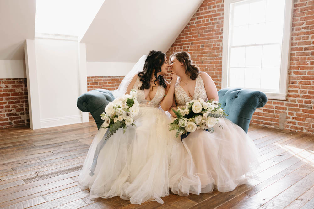 two brides in white wedding gowns with deep plunges lean toward one another to kiss 