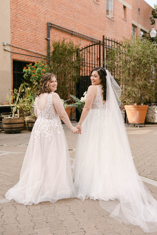 two brides hold hands and look back at the camera. they're wearing floor length wedding gowns
