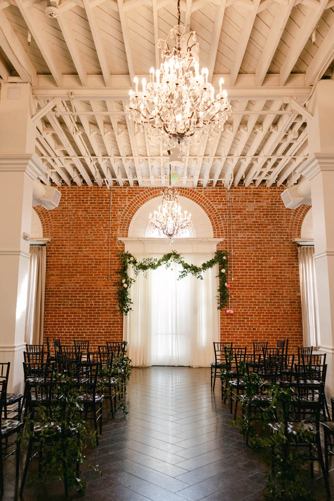 empty wedding ceremony space with brick wall, altar of green garland, a chandelier and black chairs
