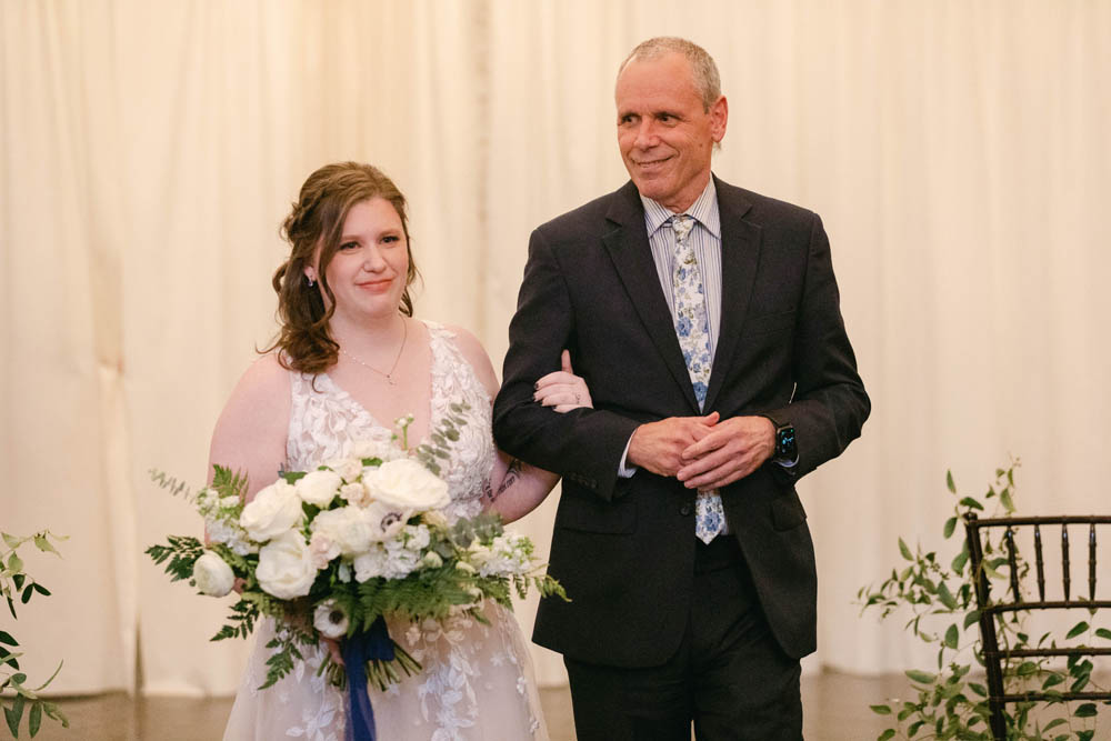 white cisgender lesbian bride walks into her LGBTQ winter wedding with a white man. she is wearing a v-neck wedding gown with lace and holding a large bouquet of white flowers with greenery. the white man is wearing a dark suit and a light blue shirt and a light blue tie.