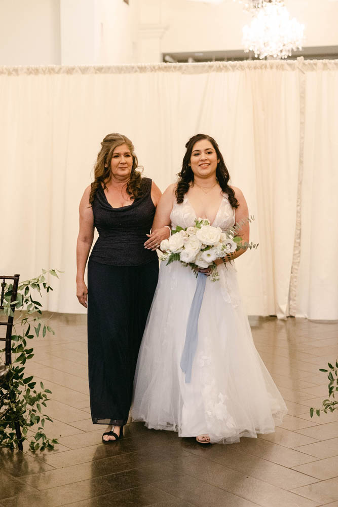 bride with dark brown hair and fair skin is escorted by a woman in a black dress