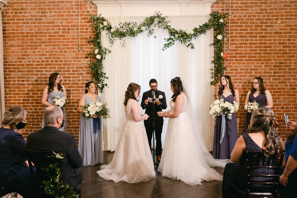 LGBTQ wedding for two brides standing at altar with an officiant in dark suit and wedding attendants wearing blue and purple dresses