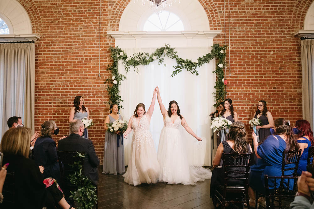 two brides hold their hands in the air after saying I do. in background is a brick wall and green garland.