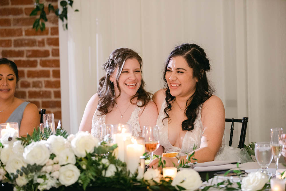 two women who were just married sit at their wedding reception smiling. both have brown hair, one lighter and one darker brown. they are smiling and laughing. 