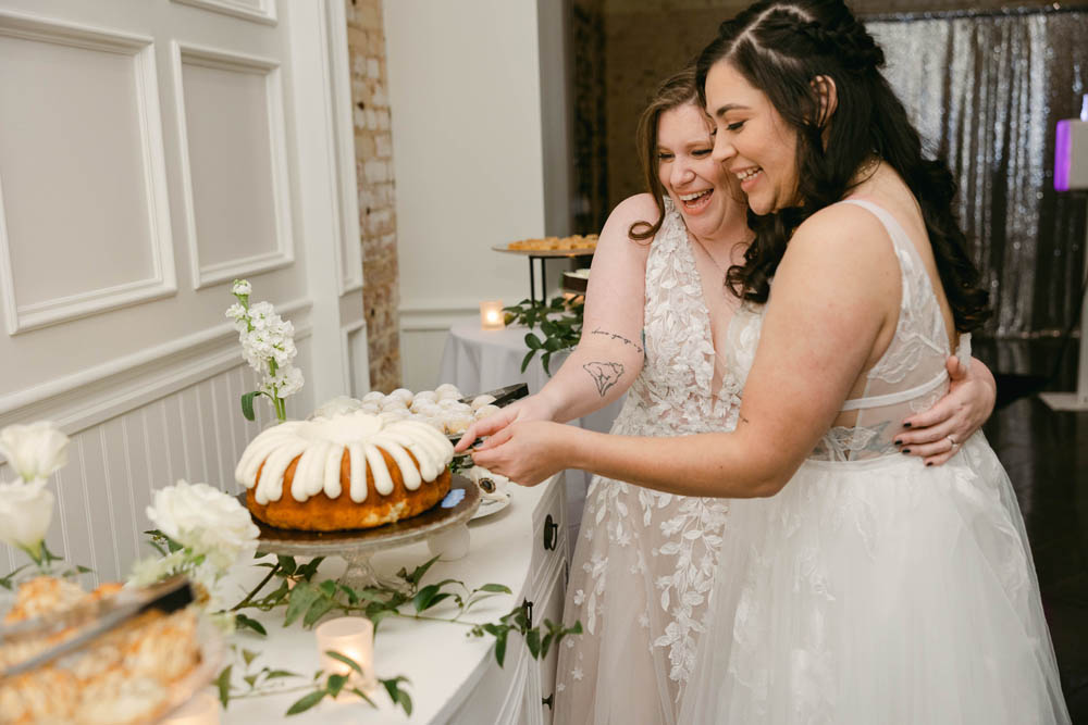 two brides in white wedding gowns cut their wedding cake