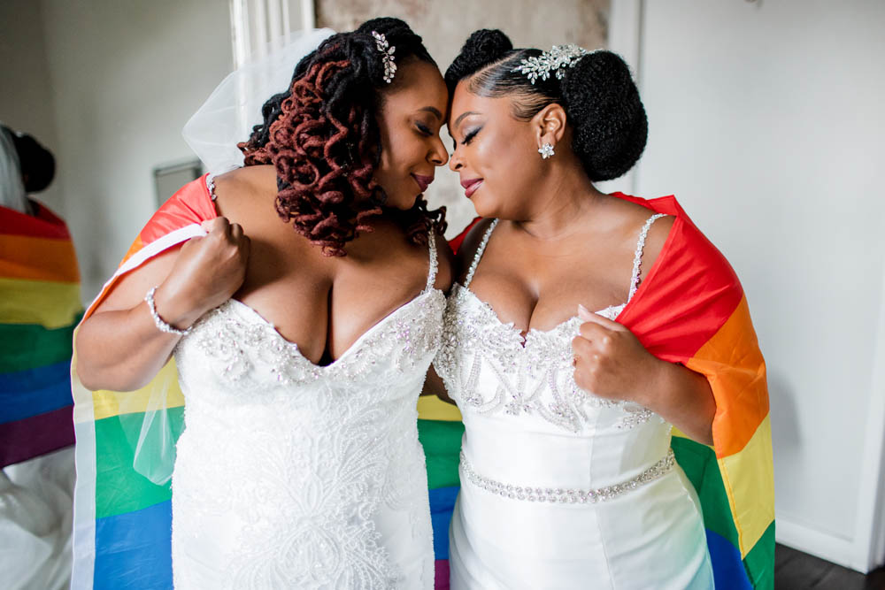 two Black brides in white wedding gowns wrap themselves in the rainbow Pride flag honoring their LGBTQ+ community