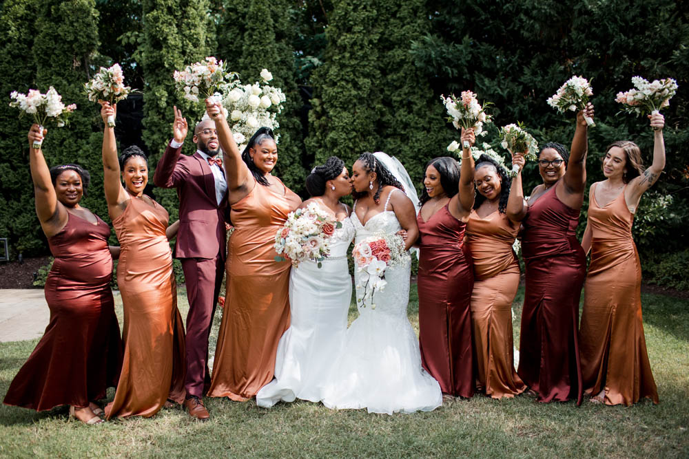 Black wedding party wearing gowns and suits in shades of burnt orange and burgundy. In the middle of the group are two Black brides wearing white floor length wedding gowns.