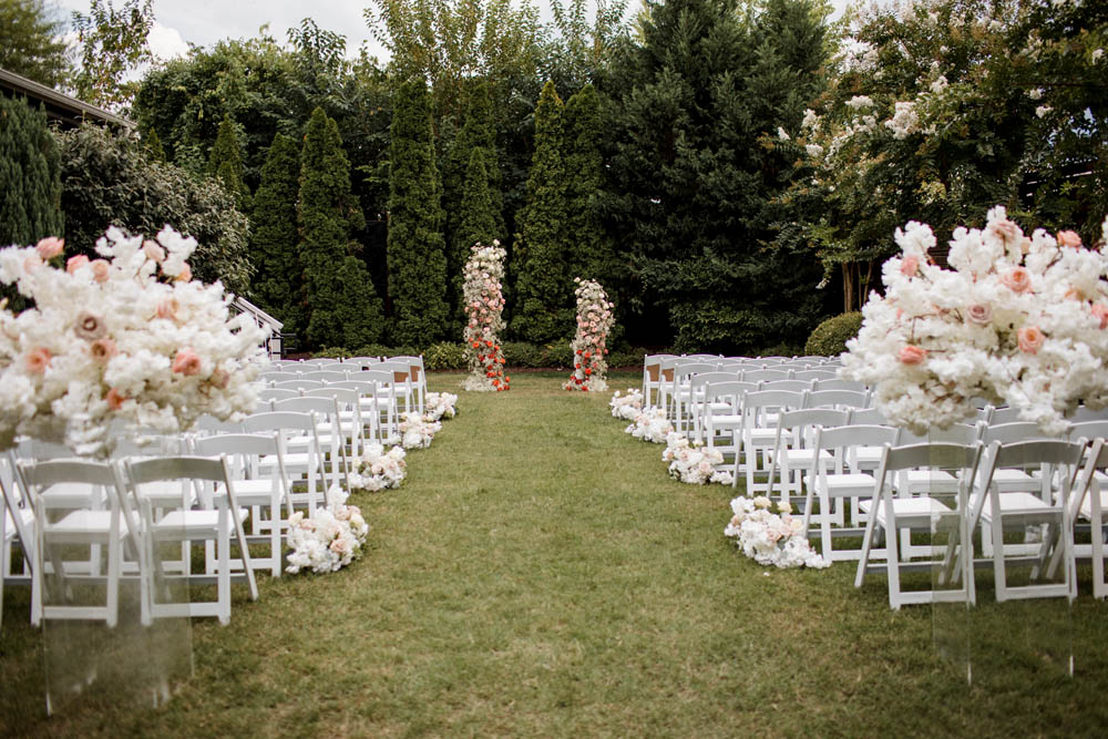 outdoor wedding with white and pink flowers and white folding chairs on green grass