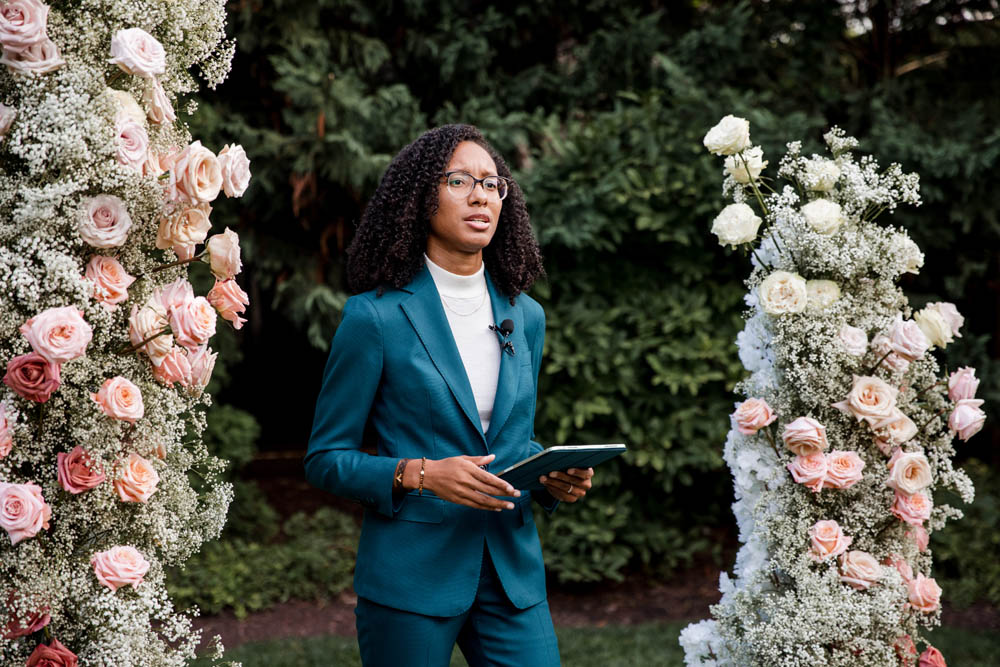 Black officiant with natural hair wearing a white mock turtleneck and marine blue suit stands between an arbor of full pink and white roses and greenery