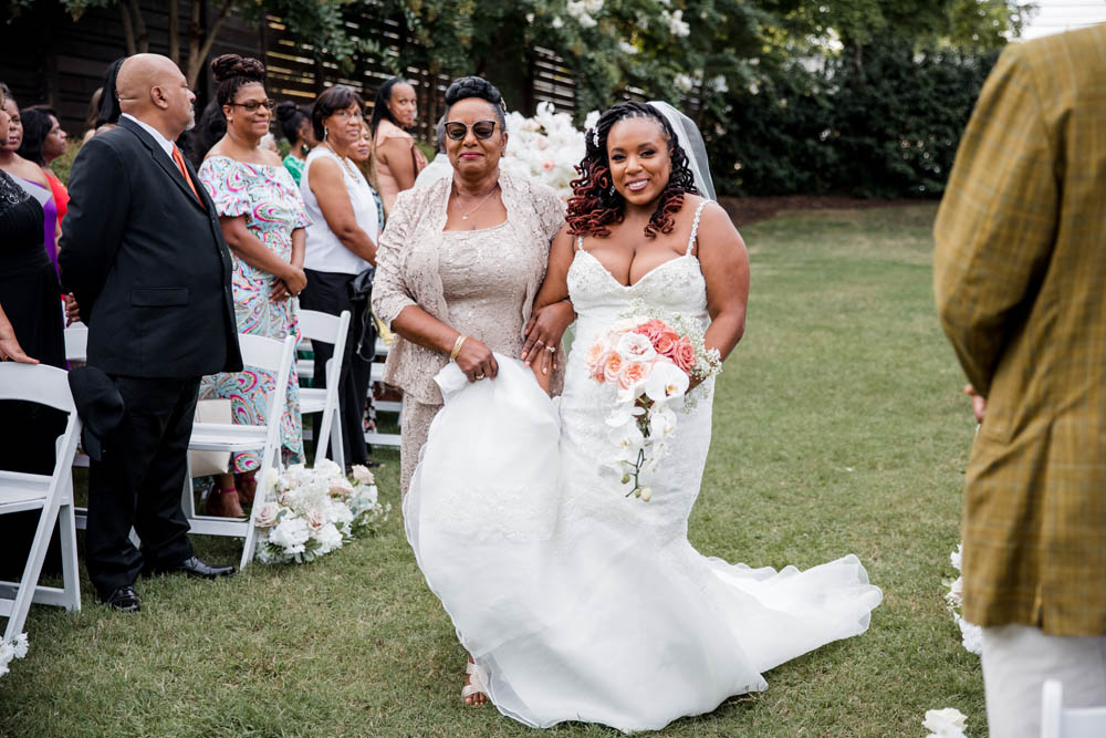 Black bride escorted down aisle with Black mother of the bride in gold dress and gold jacket and sunglasses