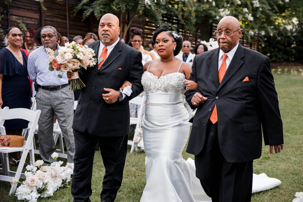 Black bride escorted by grandfather and stepfather, two Black men in black suits and wearing orange ties and orange pocket squares
