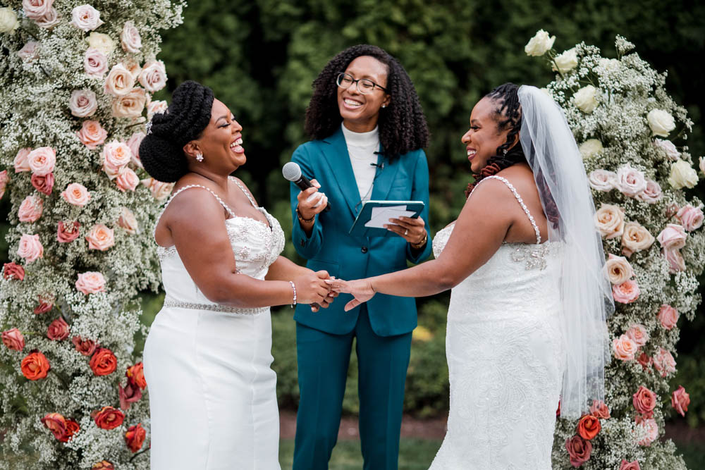 two Black brides join hands in matrimony while the officiant, a Black person with natural hair and wearing a marine blue suit smiles and points a microphone at one of the brides.