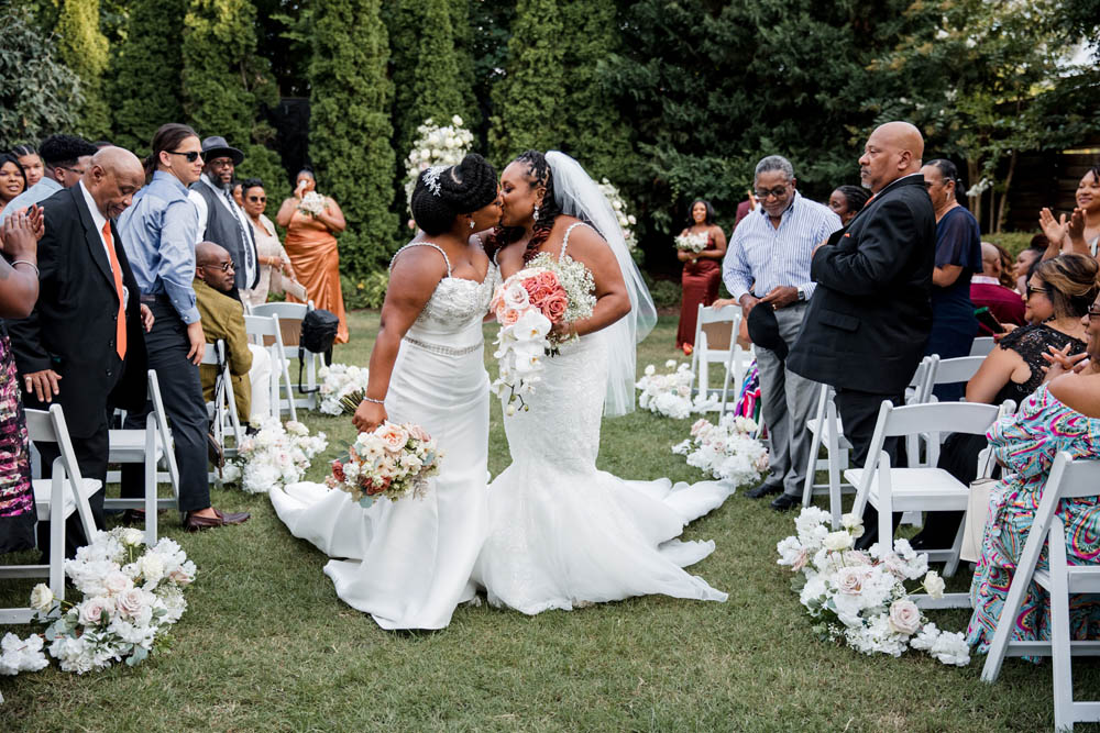 two Black brides in white wedding gowns kiss while walking down the aisle after marrying each other while guests look on