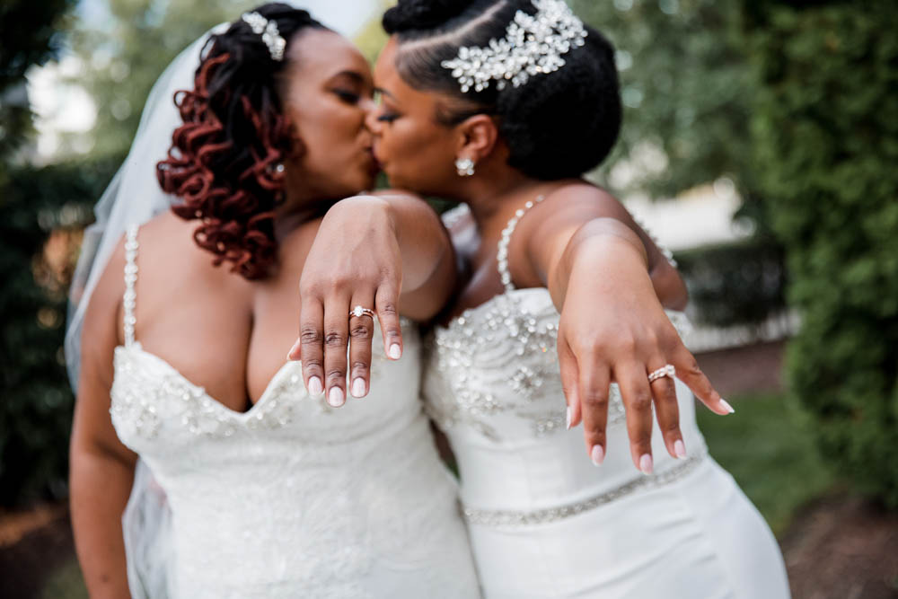 two Black brides in white wedding gowns kiss and hold out their hands showing off their wedding rings