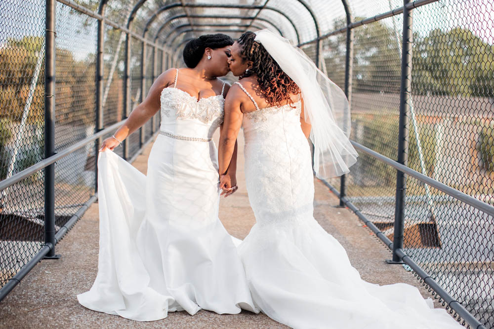 Black brides kiss while holding hands. One is facing the camera in her white wedding gown and the other is facing away from the camera showing her waist-length white veil. They're standing on a bridge covered by fencing.