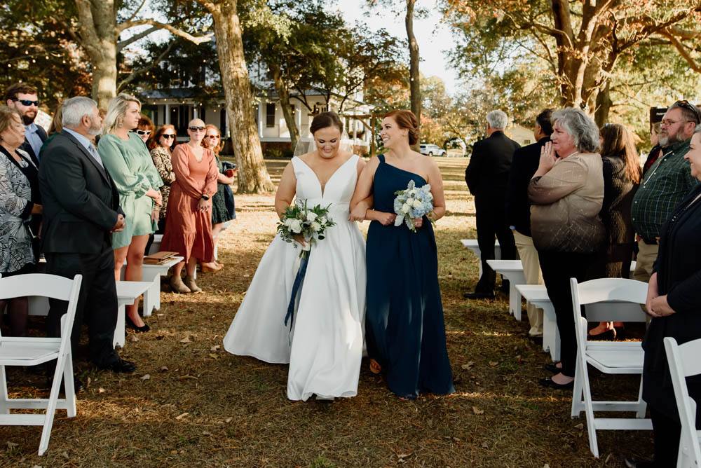 white brunette bride in white wedding gown with plunge front escorted by her matron of honor, a white woman in one-shouldered a navy gown