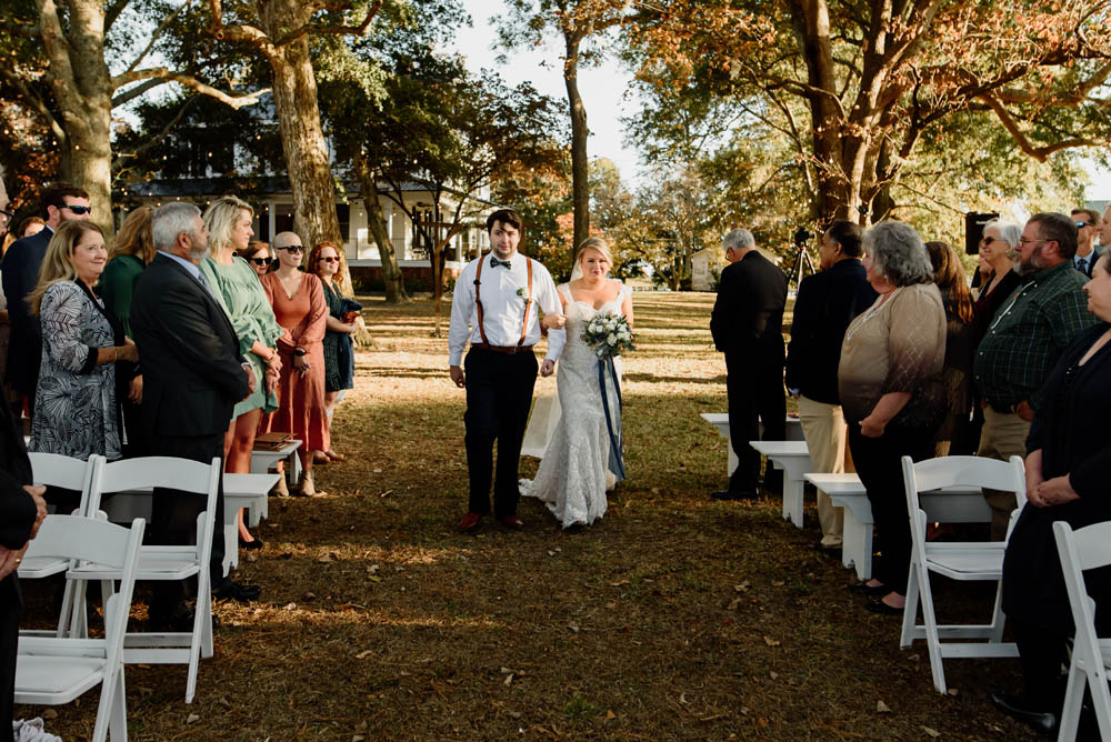 white blond bride escorted down the aisle by her brother, a white man with brown hair who is wearing a white collared shirt, brown suspenders and navy slacks as guests watch