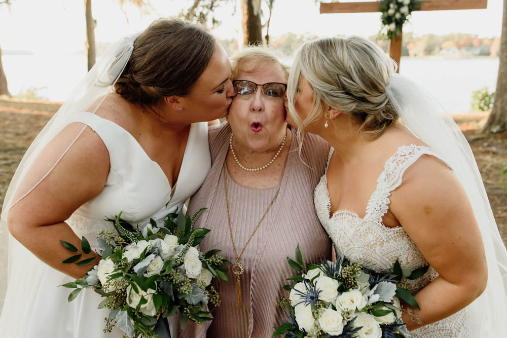 two brides kiss grandmother on each of her cheeks while she looks happy and surprised