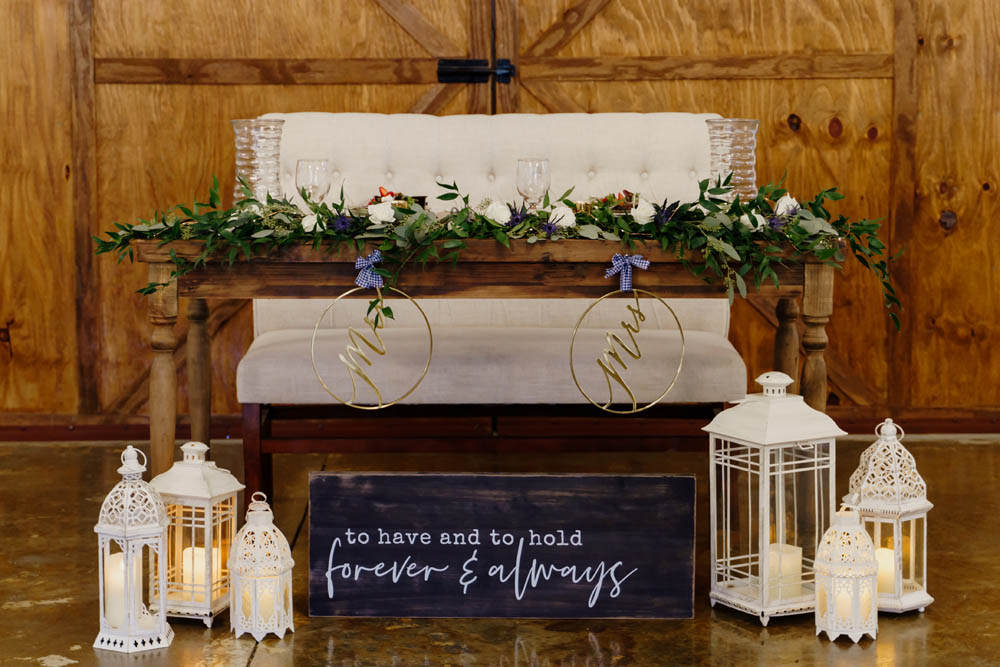 sweetheart table at lesbian Christian wedding with a bench seat, wooden table with greenery, two gold Mrs signs, white candle holders and a wooden sign painted blue with the words: To have and to hold forever and always