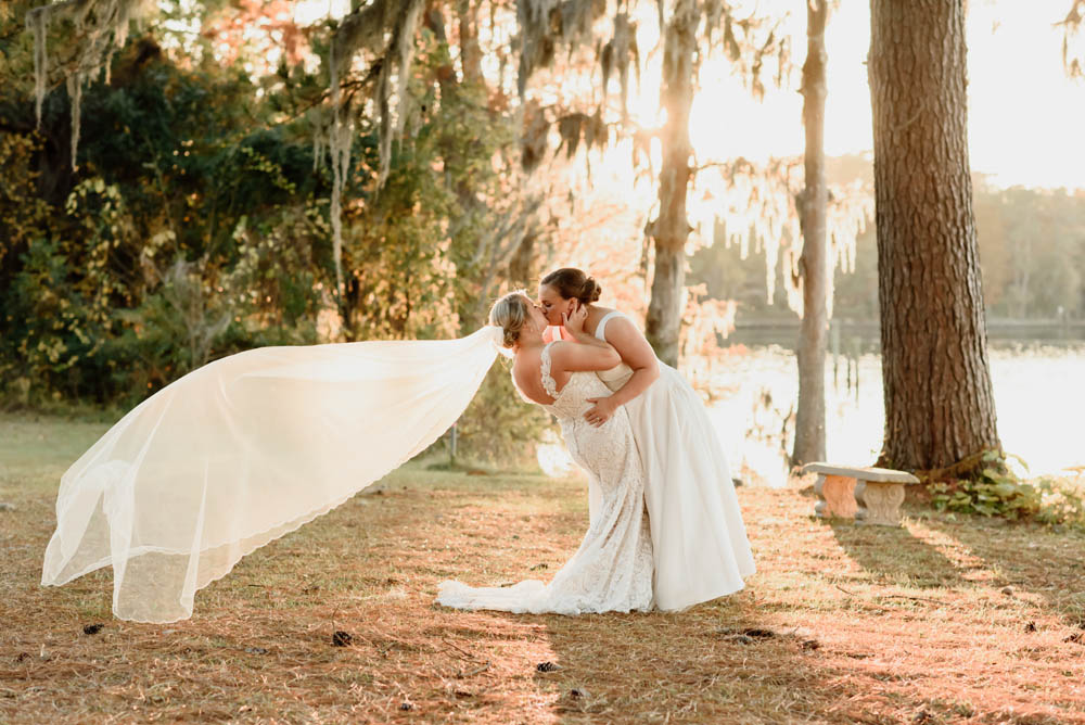 two white brides kissing outdoors by the water. one woman is leaning in and holding her wife while the one leaning back has her long veil floating in the wind