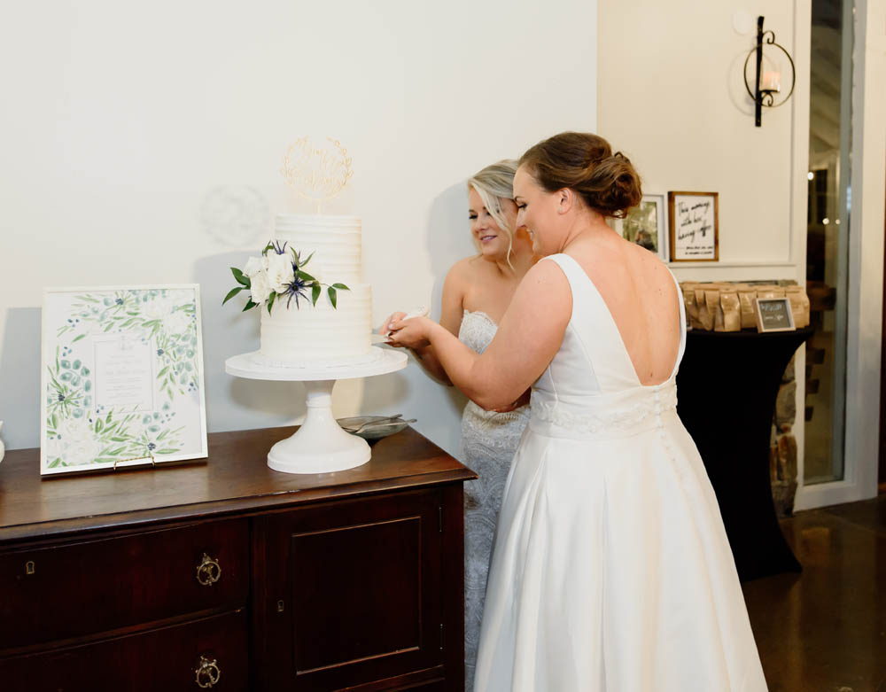 two brides in white wedding gowns cut their white wedding cake