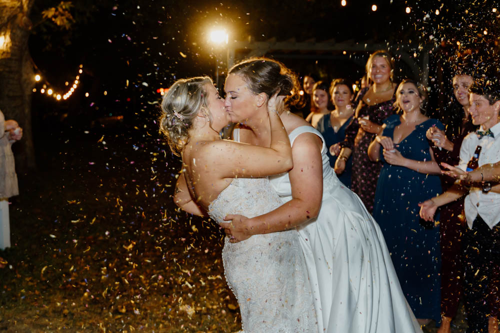 two brides kiss while dried flower confetti is tossed in their air by their wedding guests