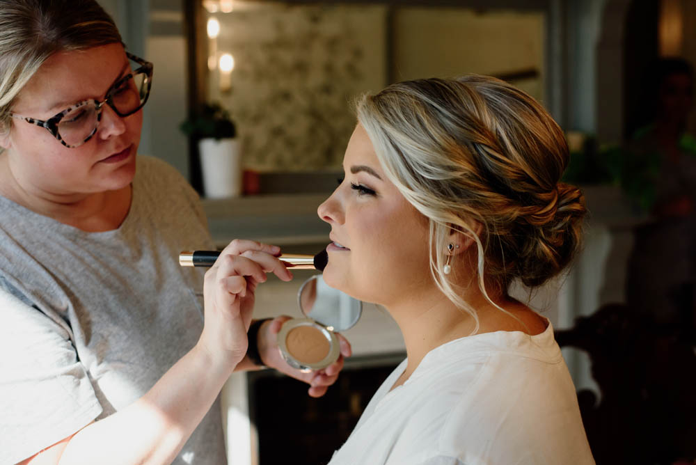 white blonde makeup artist wearing glasses applies face powder to white blonde bride with romantic up-do hairstyle