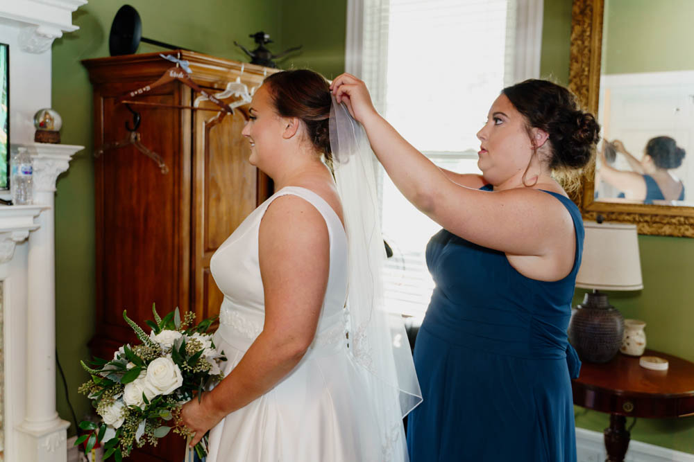 white brunette bride holding a white and green wedding bouquet has her veil put on by her bridesmaid who is a white woman with brown hair and is wearing a navy bridesmaid dress