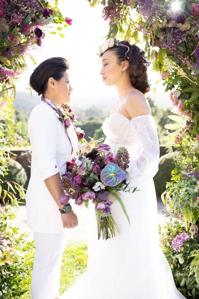 two female-presenting people in wedding attire are facing each other. One is wearing a white suit, black watch and flowers around their neck. The other is wearing an off-the-shoulder long-sleeved wedding gown and is holding a wedding bouquet of purple, pink and blue flowers.
