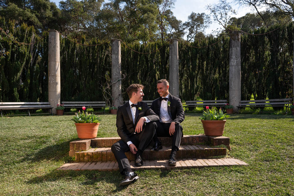 Two grooms in tuxes sit on brick steps with trees in the background on their wedding day at Filoli, an LGBTQ+ inclusive and affirming wedding venue in Woodside, California.