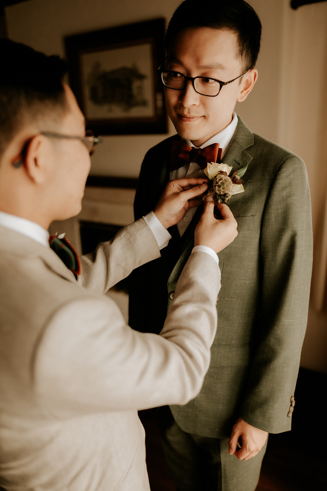 An Asian man in a tan suit pins a boutonniere on an Asian man's olive green suit. They are looking at each other with love. It's their wedding day!
