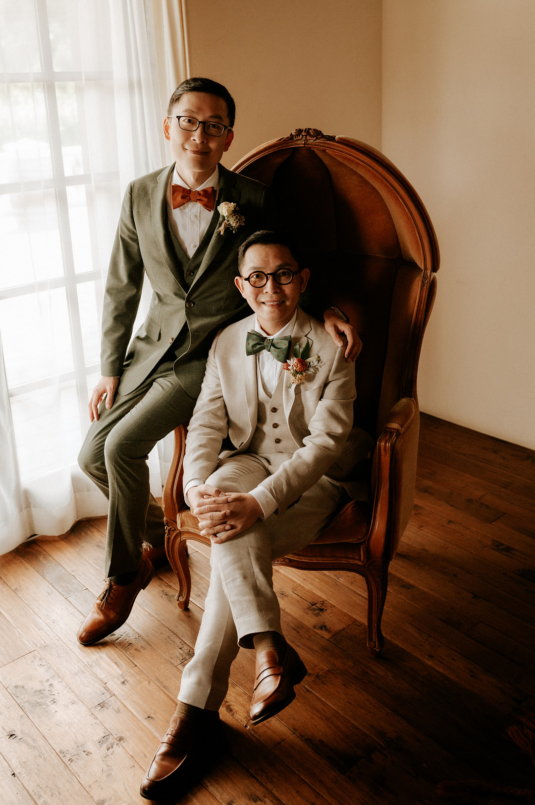 An Asian man in a tan suit and green bow tie sits in an orange chair while another Asian man in an olive green suit with an orange bow tie sits on the arm of the chair. They are both wearing glasses and are smiling at the camera.It's their wedding day!