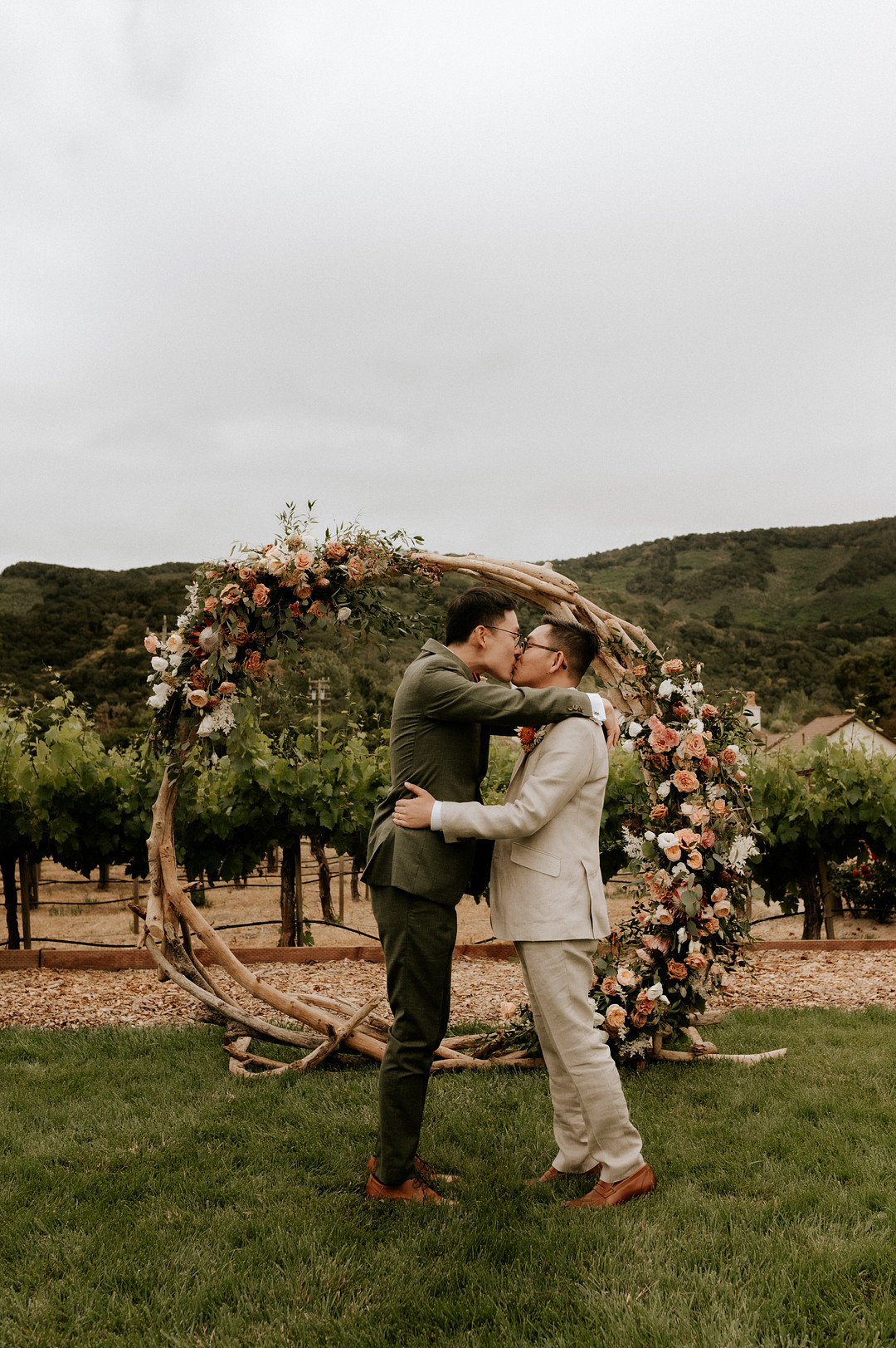 Two Asian grooms kiss in front of their flower backdrop after saying I do. One is wearing an olive green suit and the other man is wearing a cream colored suit. Mountains and a vineyard are in the background. 