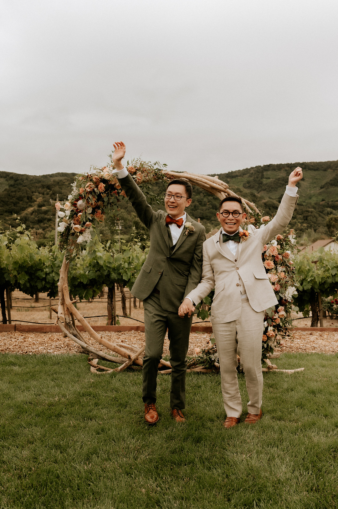 Two Asian grooms each hold up one hand in front of their flower backdrop after saying I do. One is wearing an olive green suit and the other man is wearing a cream colored suit. Mountains and a vineyard are in the background. 
