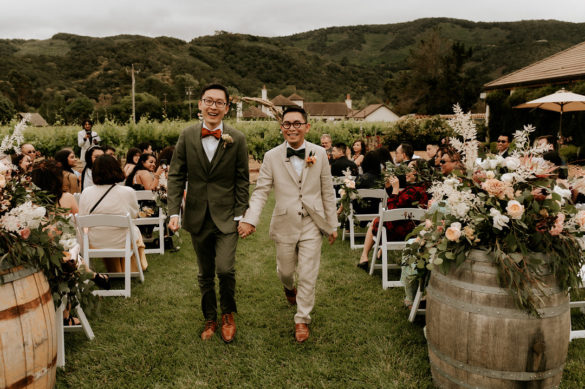 Two smiling Asian grooms in glasses walk away from their wedding guests after saying I do. One is wearing an olive green suit and an orange bow tie and the other is wearing a cream colored suit with an olive green bow tie. Mountains and a vineyard are in the background.